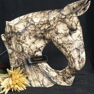 May include: A decorative horse head sculpture with a unique wood grain pattern and the name "Hondro" on a small plaque. The sculpture is cream-colored with dark brown veining, and a yellow flower is placed nearby.