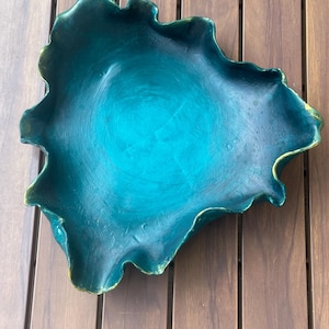 May include: A decorative bowl with an irregular, organic shape and a deep teal finish. The surface features visible brushstroke textures and subtle gold-toned highlights along the undulating, ruffled edges, creating a distinct contrast against the dark teal center.