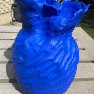 Puede incluir: Un jarrón de color azul vibrante con una superficie muy texturizada y ondulada, y un borde ancho y fruncido. El exterior presenta surcos profundos similares a tallas que crean un patrón rítmico de ondas en todo el cuerpo del recipiente.