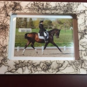 May include: A white picture frame with a black marbled design. The frame holds a photo of a brown and white horse being ridden in a dressage competition.