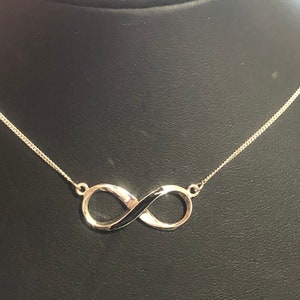 May include: A silver infinity necklace with a black enamel centre. The necklace is on a black background.