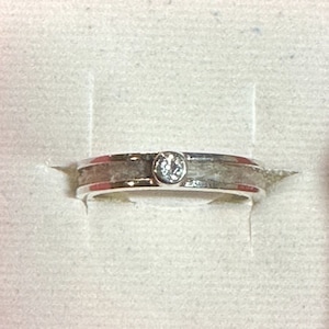 May include: A silver ring featuring a single, clear gemstone. The ring has a textured band and a polished edge. The gemstone is set in a circular bezel. The ring is displayed on a white surface.