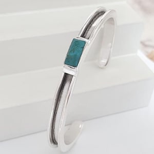 May include: A silver cuff bracelet with a rectangular turquoise stone set in the center. The bracelet has a double-band design with a smooth, polished finish. The turquoise stone adds a pop of color to the otherwise minimalist design.