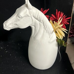 May include: A white ceramic horse head vase with a curved neck and a rounded base. The vase is filled with red and yellow flowers. The horse head is detailed with a mane and facial features.