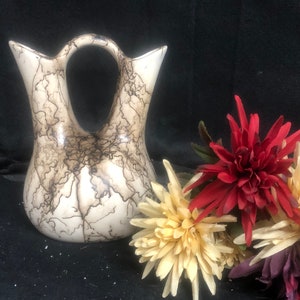 May include: A white ceramic vase with a brown crackle glaze and two spouts. The vase has a unique handle and is decorated with a natural, organic pattern. The vase is sitting on a black surface with three artificial flowers.
