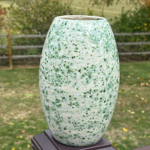 May include: A speckled ceramic vase with a light green and white glaze. The vase has a rounded body and a slightly flared opening. The exterior is covered in small, dark green spots, creating a textured appearance. The vase is displayed outdoors.