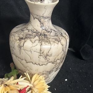 May include: A cream-coloured vase with a lid, displaying a distinctive black lightning pattern. The vase has a rounded body and a flared neck. Artificial flowers in red and yellow are in the foreground. The vase is likely ceramic or porcelain.