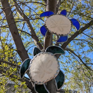 May include: Two stained glass turtle ornaments with white sand dollar centers and blue and green flipper accents hang from a tree branch. The ornaments are framed with silver-colored metal.
