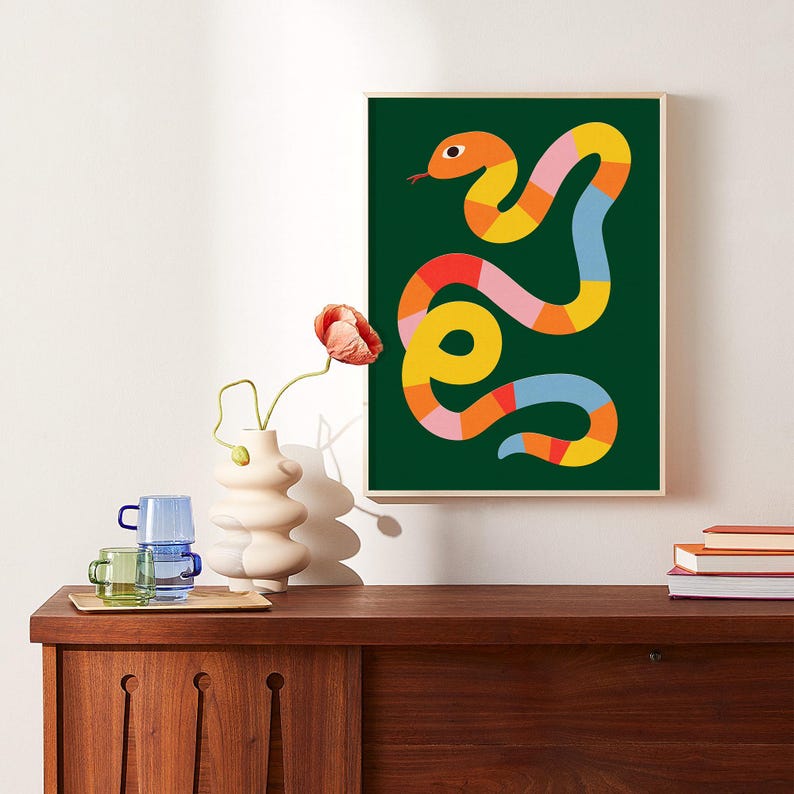 May include: An abstract illustration of a snake on a green background. The snake is made up of yellow, orange, red, pink, and blue segments. The illustration is framed in a light wood frame.