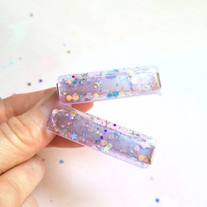 May include: Two clear purple rectangular hair clips with iridescent glitter and stars inside.