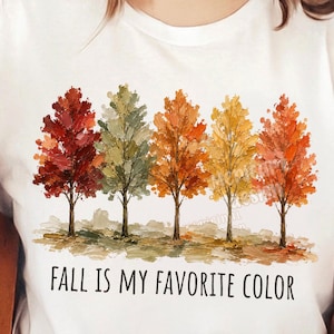 K&ouml;nnte beinhalten: Ein wei&szlig;es kurz&auml;rmeliges T-Shirt mit einer Grafik von f&uuml;nf stilisierten B&auml;umen in herbstlichen Farben wie Rot, Gr&uuml;n, Orange und Gelb. Unter den B&auml;umen steht der Text FALL IS MY FAVORITE COLOR in schwarzen Gro&szlig;buchstaben.