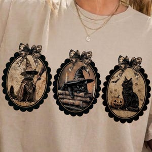 May include: A beige short-sleeve t-shirt featuring three oval-shaped graphic prints. Each oval is framed by a black scalloped border and a bow at the top, depicting black cats in witch-themed settings with books, a broom, and a pumpkin.