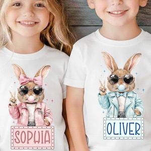 May include: Two white t-shirts with cartoon bunny graphics. One bunny wears a pink jacket and bow, the other a blue jacket and sunglasses. Each holds up a peace sign and has a nameplate: "SOPHIA" and "OLIVER".