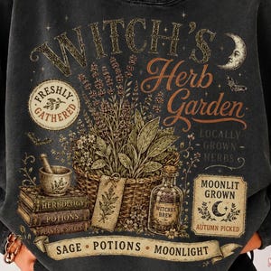 May include: A charcoal gray crewneck sweatshirt featuring a graphic print titled Witch's Herb Garden. The design includes a woven basket of herbs, a mortar and pestle, stacked books, a glass bottle, and text reading Freshly Gathered and Moonlit Grown.