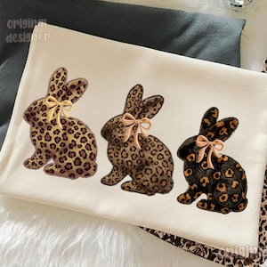 May include: Three decorative bunny rabbits with leopard print patterns and bows. The bunnies are in shades of brown and black, with tan bows. The background is a cream-colored fabric.
