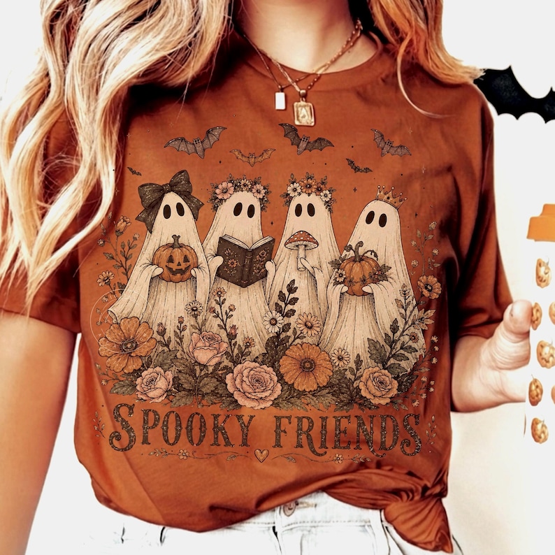 May include: A rust-colored t-shirt featuring a graphic of four ghosts surrounded by flowers and bats. The ghosts hold a pumpkin, a book, and a mushroom. The text "SPOOKY FRIENDS" is printed at the bottom in a dark, decorative font style.