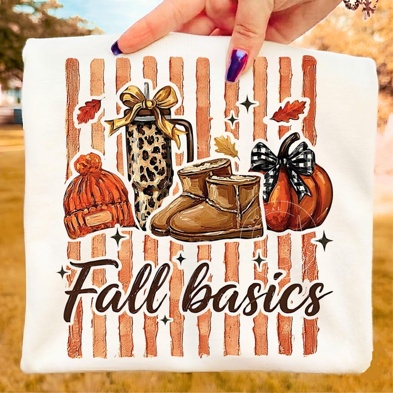 May include: A white t-shirt featuring a graphic of vertical orange stripes and the text Fall basics. The design includes an orange knit beanie, a leopard print tumbler with a gold bow, brown boots, and an orange pumpkin with a black plaid bow.