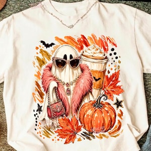 Puede incluir: Una camiseta blanca con un gr&aacute;fico de un fantasma que lleva una estola de piel rosa, gafas de sol de ojo de gato y un collar de joyas. El fantasma sostiene un bolso rosa junto a una calabaza, una taza de caf&eacute; y hojas de arce naranjas.