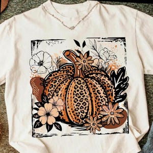 May include: A cream-colored t-shirt featuring a central graphic of a pumpkin with a brown leopard print pattern. The pumpkin is surrounded by black and white line-art flowers, leaves, and abstract brown shapes, all enclosed within a square frame.