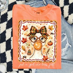 May include: A peach-colored t-shirt featuring a graphic of a goose wearing a plaid bow and heart-shaped glasses while blowing a bubble. The design includes autumn leaves, pumpkins, and the text "just a girl who loves fall" in white script.