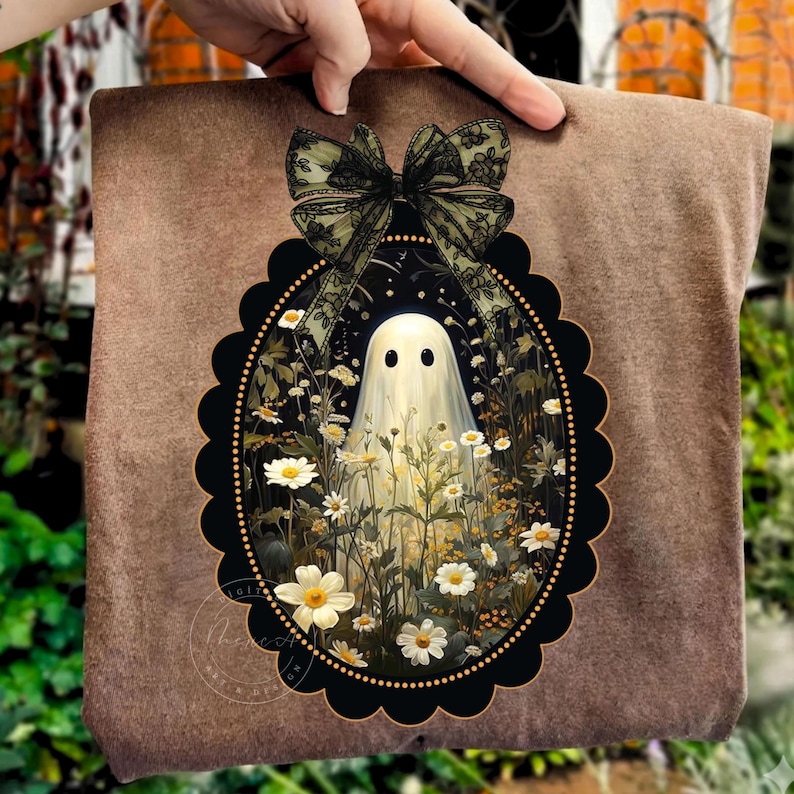 May include: A brown t-shirt featuring a central oval graphic with a scalloped black border. The design depicts a white ghost surrounded by white and yellow wildflowers, topped with a decorative sheer black lace bow with a floral pattern.