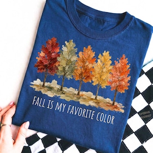 K&ouml;nnte beinhalten: Ein marineblaues T-Shirt mit einer Grafik von f&uuml;nf B&auml;umen mit strukturierten, malerischen Bl&auml;ttern in Rot-, Gr&uuml;n-, Orange- und Gelbt&ouml;nen. Unter den B&auml;umen steht der Text "FALL IS MY FAVORITE COLOR" in einer einfachen, wei&szlig;en, serifenlosen Schrift.