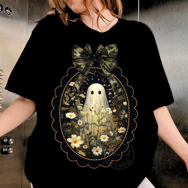 May include: A black t-shirt featuring a central graphic of a white ghost standing in a field of white daisies. The design is enclosed in a scalloped oval frame with a large, dark green lace-style bow positioned at the top of the oval border.