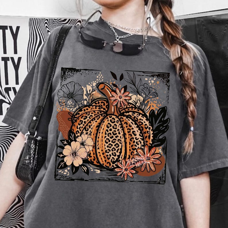 May include: A charcoal gray short-sleeve t-shirt featuring a central graphic of a pumpkin with a leopard print pattern. The pumpkin is surrounded by stylized flowers and leaves in shades of orange, brown, and black, all enclosed in a square frame.