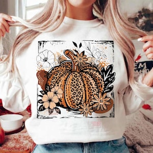 May include: A white long-sleeve sweatshirt featuring a central graphic of a pumpkin with a leopard print pattern. The pumpkin is surrounded by black and tan line-art flowers and leaves, all enclosed within a distressed black square border.