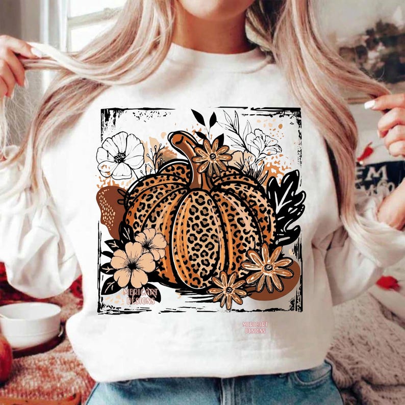 May include: A white long-sleeve sweatshirt featuring a central graphic of a pumpkin with a leopard print pattern. The pumpkin is surrounded by black and tan line-art flowers and leaves, all enclosed within a distressed black square border.