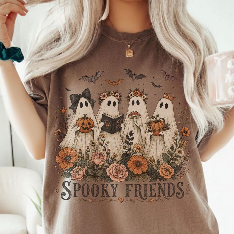 May include: A brown t-shirt featuring a graphic of four ghosts adorned with flowers, a bow, a crown, and a book. The ghosts are surrounded by orange and pink flowers, bats, and pumpkins. The text "SPOOKY FRIENDS" is printed in black at the bottom.