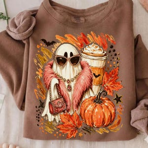 Puede incluir: Una sudadera marr&oacute;n de cuello redondo con un gr&aacute;fico de un fantasma con gafas de sol, una estola de piel sint&eacute;tica rosa y un bolso. El dise&ntilde;o incluye un caf&eacute; con nata, una calabaza naranja y hojas de oto&ntilde;o en tonos rojos y anaranjados.