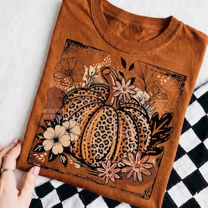 May include: A rust-colored t-shirt featuring a central graphic of a pumpkin with a leopard print pattern. The pumpkin is surrounded by various line-art flowers and leaves within a rectangular frame. The text MERICART DESIGNS is visible.