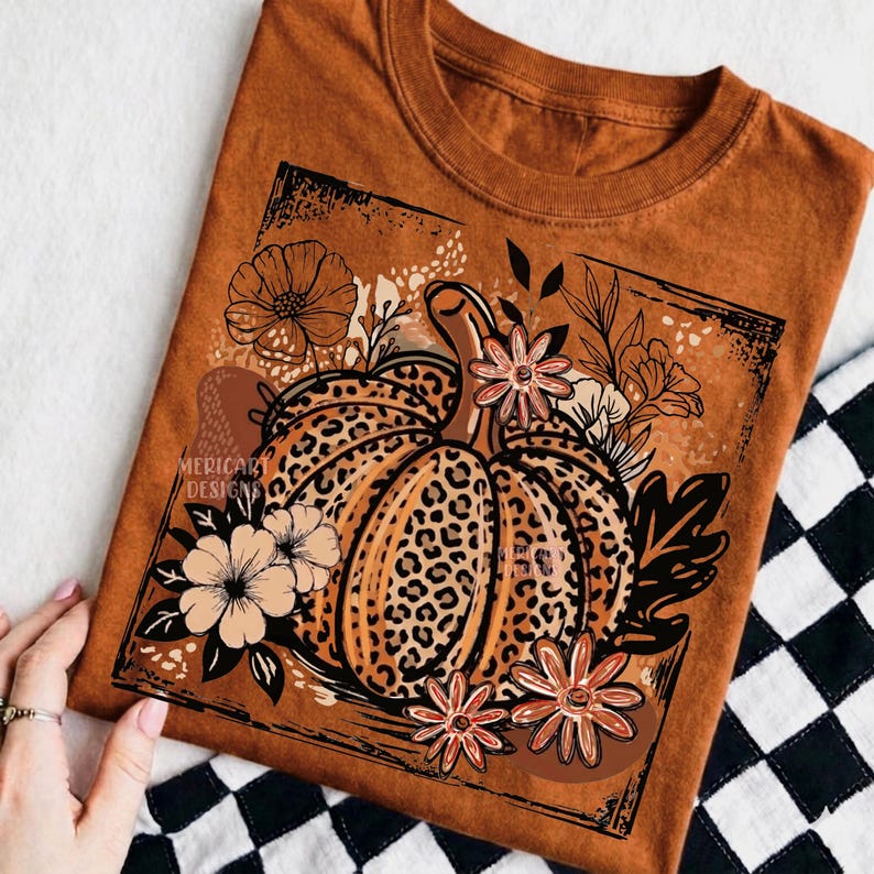 May include: A rust-colored t-shirt featuring a central graphic of a pumpkin with a leopard print pattern. The pumpkin is surrounded by various line-art flowers and leaves within a rectangular frame. The text MERICART DESIGNS is visible.