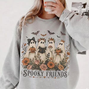 May include: A heather gray crewneck sweatshirt featuring a graphic of four ghosts adorned with floral crowns, holding a pumpkin, a book, and a mushroom. The design includes bats and the text "SPOOKY FRIENDS" in a dark, decorative serif font.