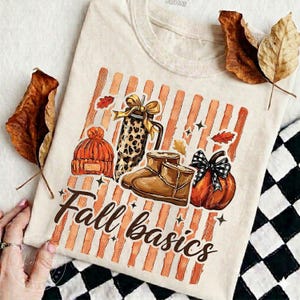 May include: A cream-colored t-shirt featuring a graphic of orange vertical stripes, an orange knit hat, a leopard print tumbler, brown boots, and an orange pumpkin with a black and white plaid bow. The text "Fall basics" is printed in brown script.