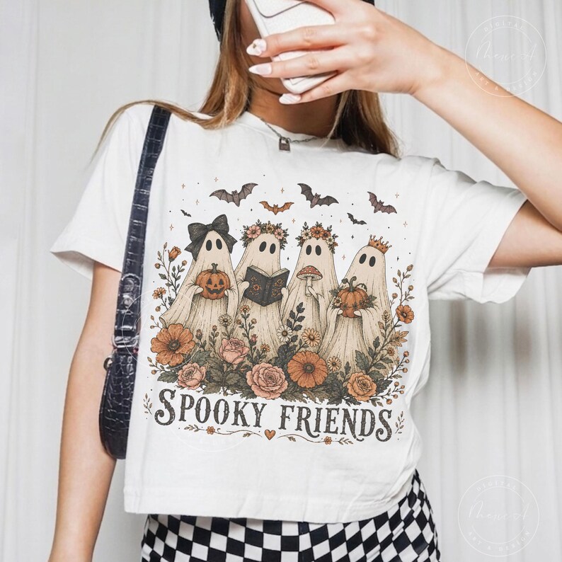 May include: A white t-shirt featuring a graphic print of four ghosts surrounded by flowers and bats. The ghosts are holding a pumpkin, a book, and a mushroom. The text "SPOOKY FRIENDS" is printed in a dark, decorative font at the bottom of the design.