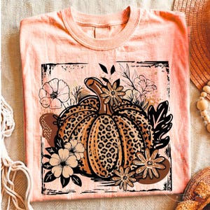 May include: A peach-colored t-shirt featuring a central graphic of a pumpkin with a leopard print pattern. The pumpkin is surrounded by black and white line-art flowers and leaves, all contained within a distressed, square-shaped black border.