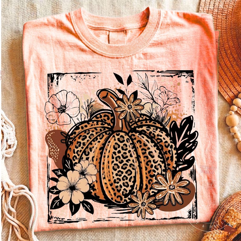 May include: A peach-colored t-shirt featuring a central graphic of a pumpkin with a leopard print pattern. The pumpkin is surrounded by black and white line-art flowers and leaves, all contained within a distressed, square-shaped black border.