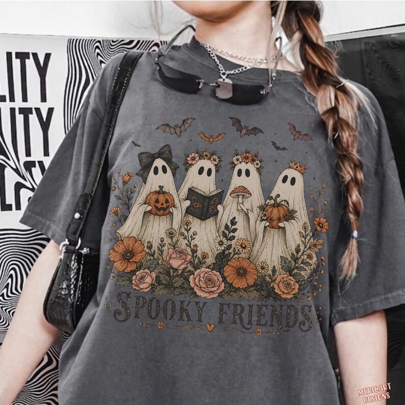 May include: A charcoal gray t-shirt featuring a graphic print of four ghosts surrounded by orange and pink flowers. The ghosts hold a pumpkin, a book, a mushroom, and a pumpkin respectively. The text "SPOOKY FRIENDS" is printed below the ghosts.