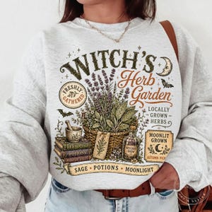 May include: A light gray crewneck sweatshirt featuring a graphic print with the text Witch's Herb Garden, Freshly Gathered, Locally Grown Herbs, Moonlit Grown, and Sage Potions Moonlight. The design includes a basket of herbs, books, and a bottle.