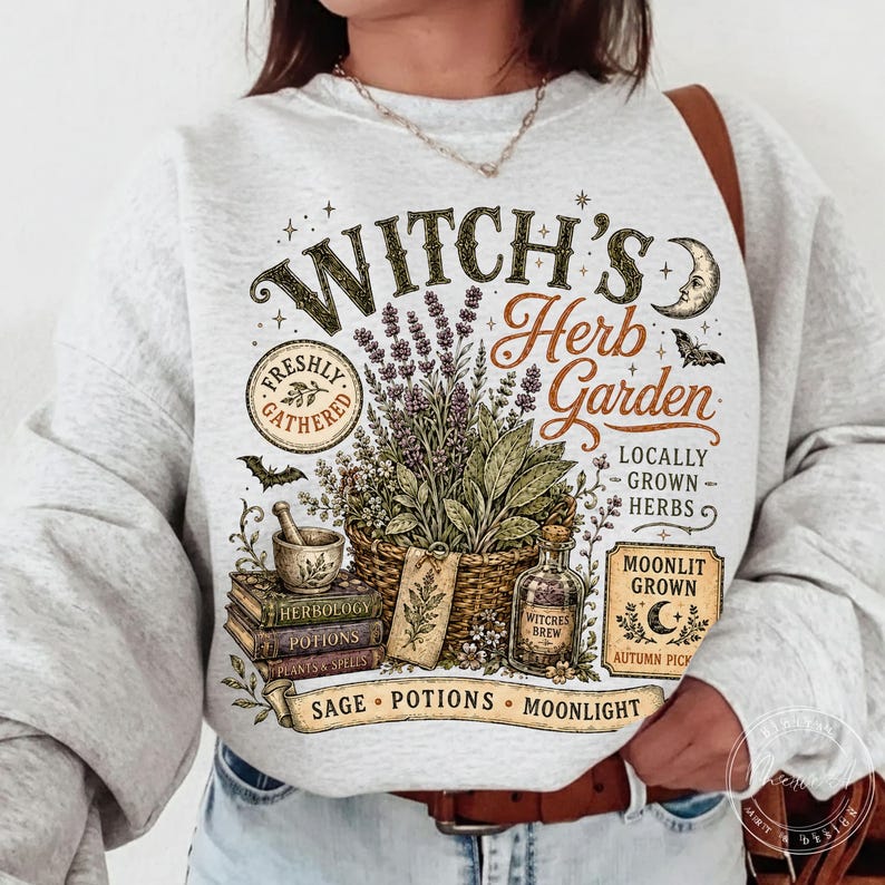 May include: A light gray crewneck sweatshirt featuring a graphic print with the text Witch's Herb Garden, Freshly Gathered, Locally Grown Herbs, Moonlit Grown, and Sage Potions Moonlight. The design includes a basket of herbs, books, and a bottle.
