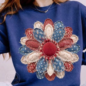 Puede incluir: Una sudadera azul marino con un gran diseño de flores aplicado. Los pétalos de la flor son una mezcla de telas rojas, blancas y azules con varios patrones. El centro de la flor es un círculo rojo liso. El texto "AMERICAN DESIGN" es visible.