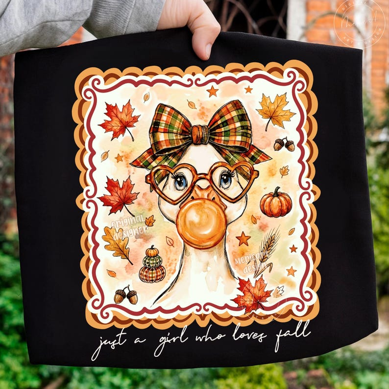 May include: A black t-shirt featuring a graphic of a goose wearing a plaid bow and heart-shaped glasses while blowing an orange bubble. The design includes autumn leaves, pumpkins, and acorns, with the text "just a girl who loves fall" below.