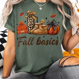 May include: A dark olive green t-shirt featuring a graphic print of a leopard print tumbler with a gold bow, a pair of tan boots, an orange knit beanie, and a pumpkin with a black and white plaid bow. The text Fall basics is printed below.