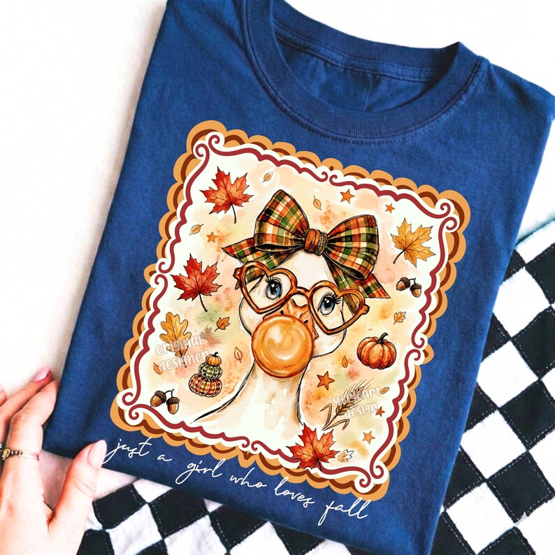 May include: A navy blue t-shirt featuring a graphic of a goose wearing a plaid bow and heart-shaped glasses while blowing a bubble. The design includes autumn leaves, pumpkins, and acorns, with the text "just a girl who loves fall" below.