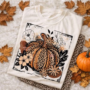 May include: A cream-colored t-shirt featuring a rectangular graphic of a pumpkin with a brown leopard print pattern. The pumpkin is surrounded by black and white line-art flowers and leaves, with brown abstract shapes and the text MERICART DESIGNS.