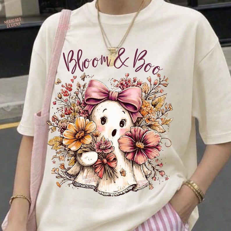 May include: A cream-colored t-shirt featuring a graphic print of a ghost with a pink bow, surrounded by orange and pink flowers. The text "Bloom & Boo" is printed in a dark script font above the ghost. The shirt has short sleeves and a crew neck.