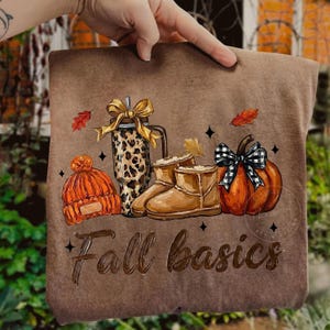 May include: A brown t-shirt featuring a graphic print of an orange knit beanie, a leopard print tumbler with a gold bow, tan boots, and an orange pumpkin with a black and white plaid bow. The text Fall basics is printed in brown script below.