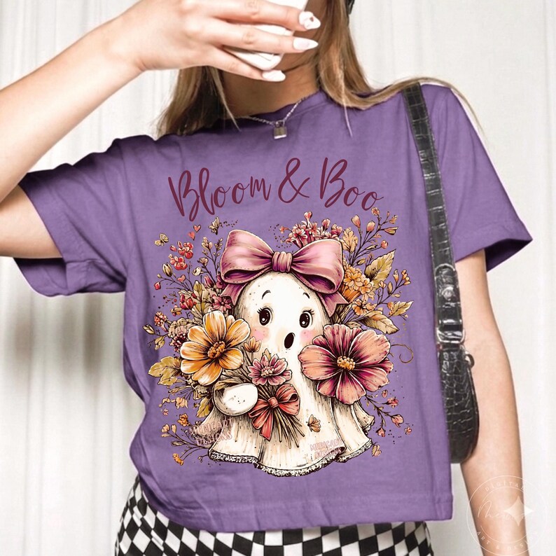 May include: A purple t-shirt featuring a graphic of a white ghost wearing a pink bow, surrounded by colorful autumn flowers and foliage. The text "Bloom & Boo" is printed in a dark script font above the ghost illustration on the front of the shirt.